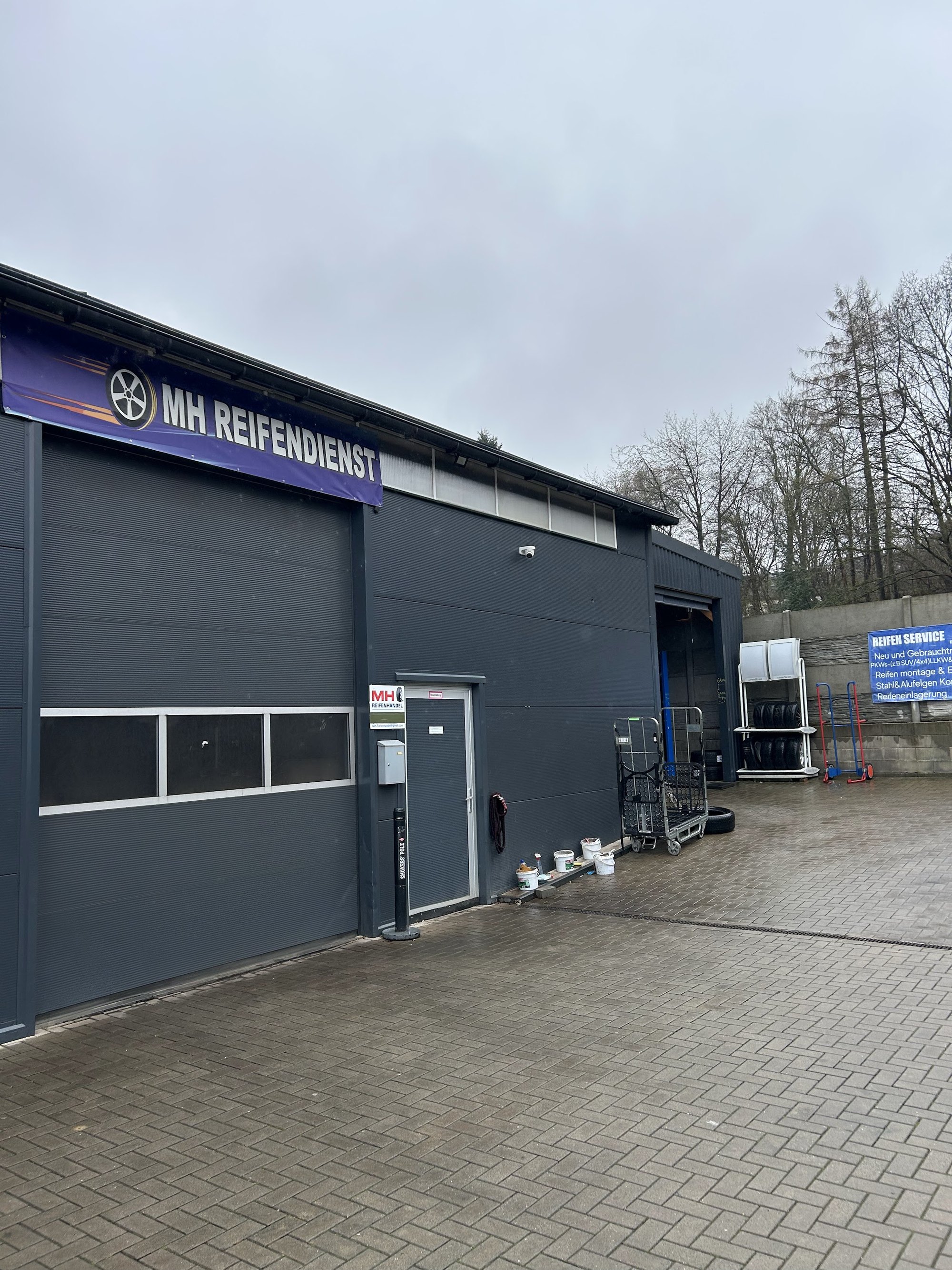 Seitenansicht der dunklen Halle von MH Reifendienst mit violettem Banner, Eingangstür, Reifenständer und gepflastertem Hof